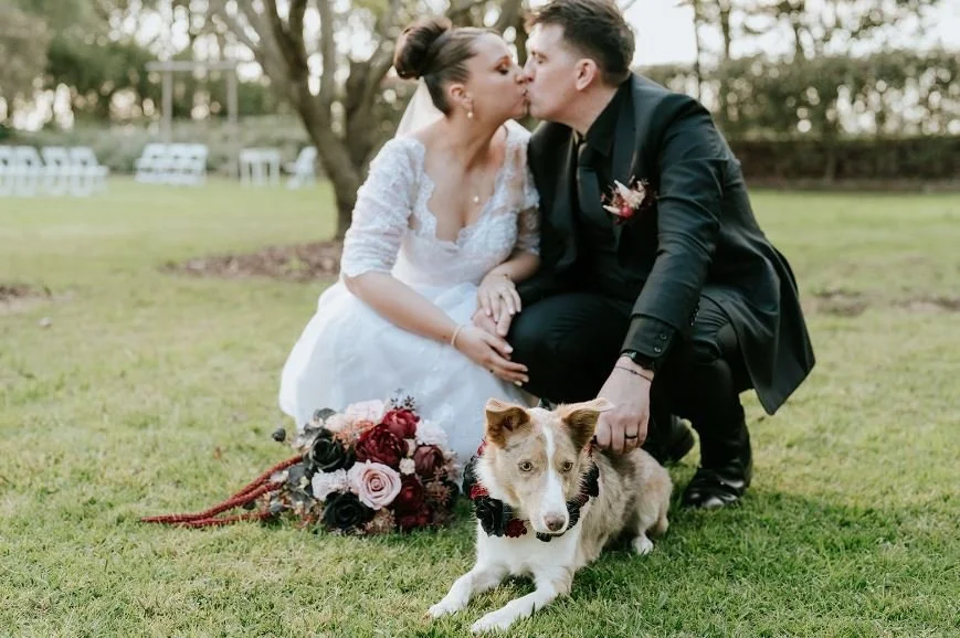 Couple in wedding attire kissing outdoors, with a dog lying on the grass in front, and a bouquet of flowers nearby.