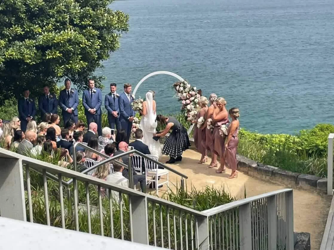 A wedding ceremony taking place outdoors near a body of water, with the bride and groom standing under an arch decorated with flowers, surrounded by bridesmaids and groomsmen, and seated guests observing.