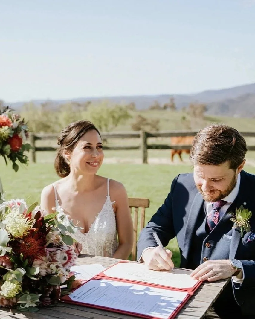 A bride with dark hair in a white dress smiling at a groom with brown hair and a beard in a navy suit, signing a document on a wooden table outdoors. There are flowers and a landscape with green fields, trees, and hills in the background.