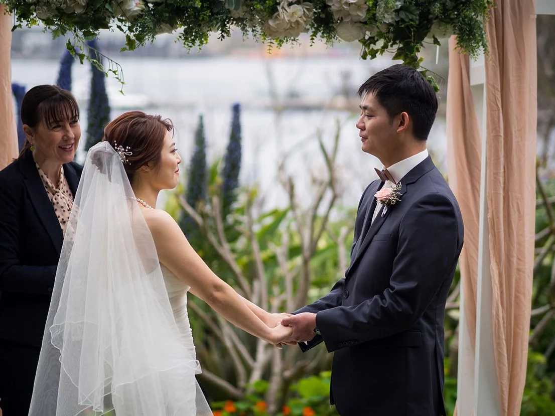A bride and groom holding hands during a wedding ceremony outdoors, with an officiant smiling nearby. Green plants and flowers decorate the backdrop.