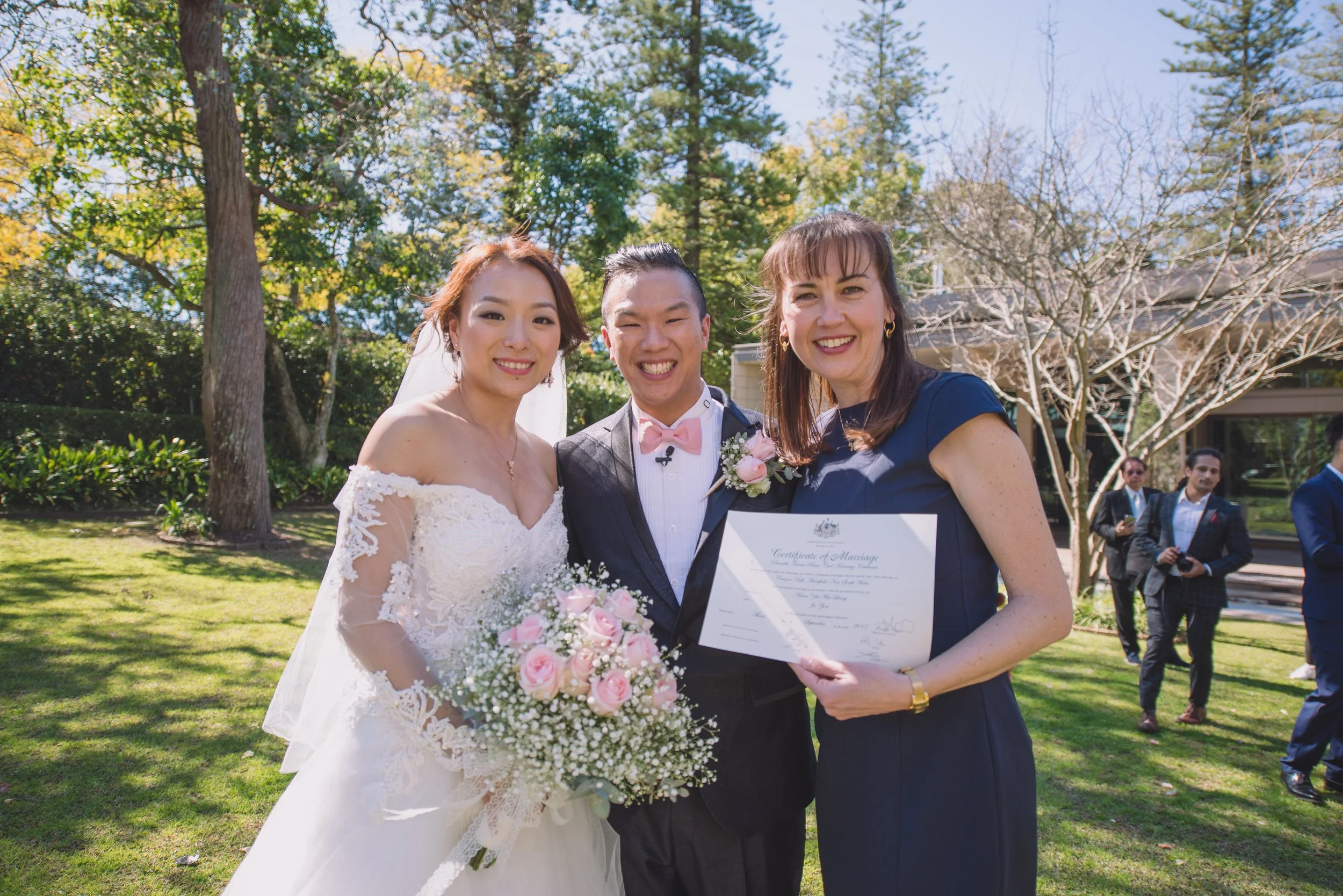 A wedding celebration outdoors with three people smiling, including a bride in a white gown holding a bouquet of pink roses, a groom in a dark suit with a pink bow tie and boutonniere, and a woman in a navy dress holding a wedding certificate, with t