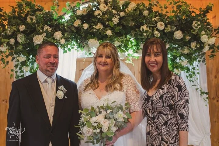 A happy bride in a lace wedding dress holding a bouquet, standing between a groom in a black suit and a woman in a patterned dress, with a flowered arch and draped fabric behind them at a wedding celebration.