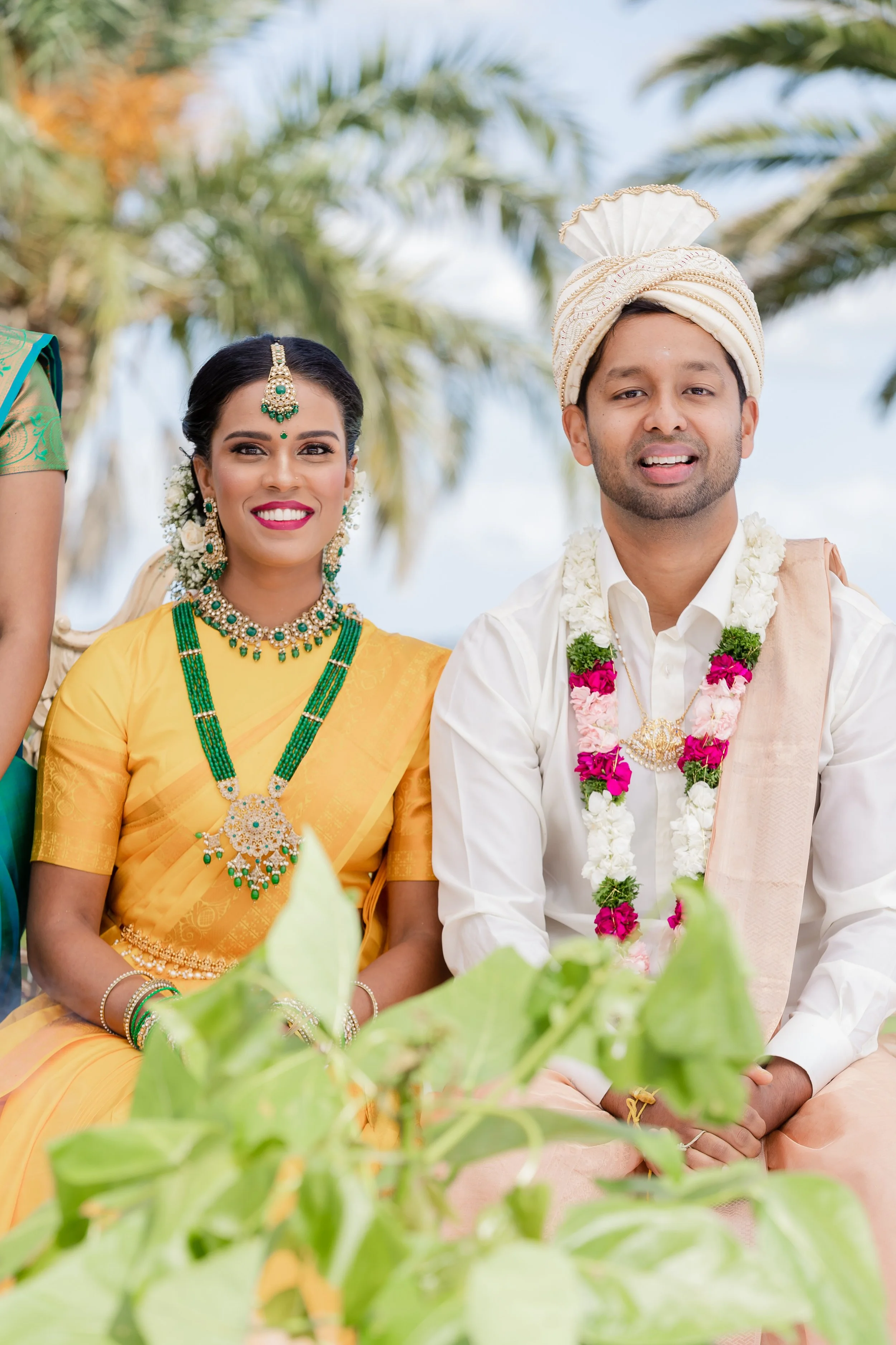 A South Asian couple dressed in traditional wedding attire outdoors, with palm trees in the background.