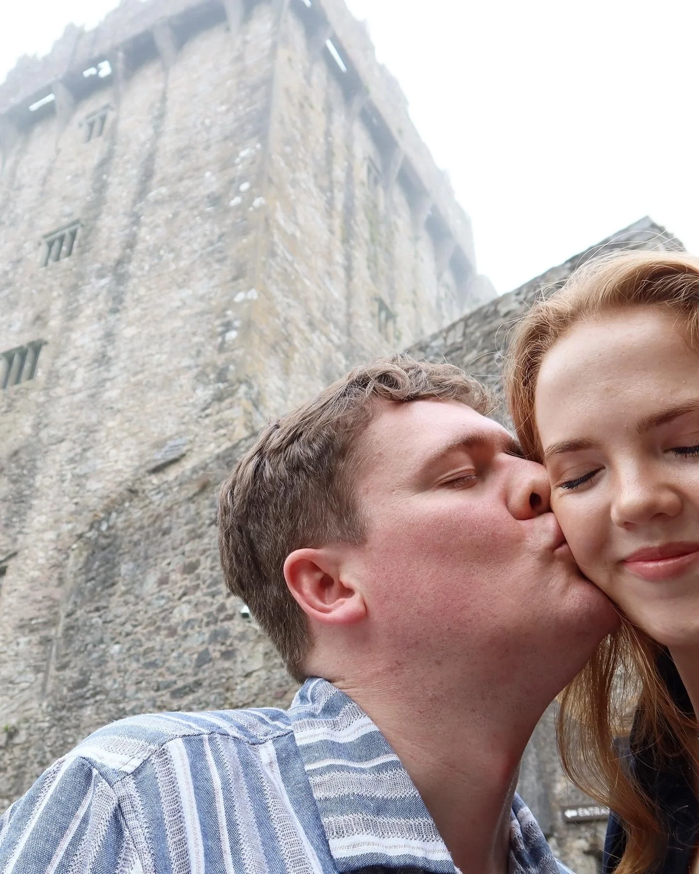 A man is kissing a woman on the cheek in front of a large stone castle.