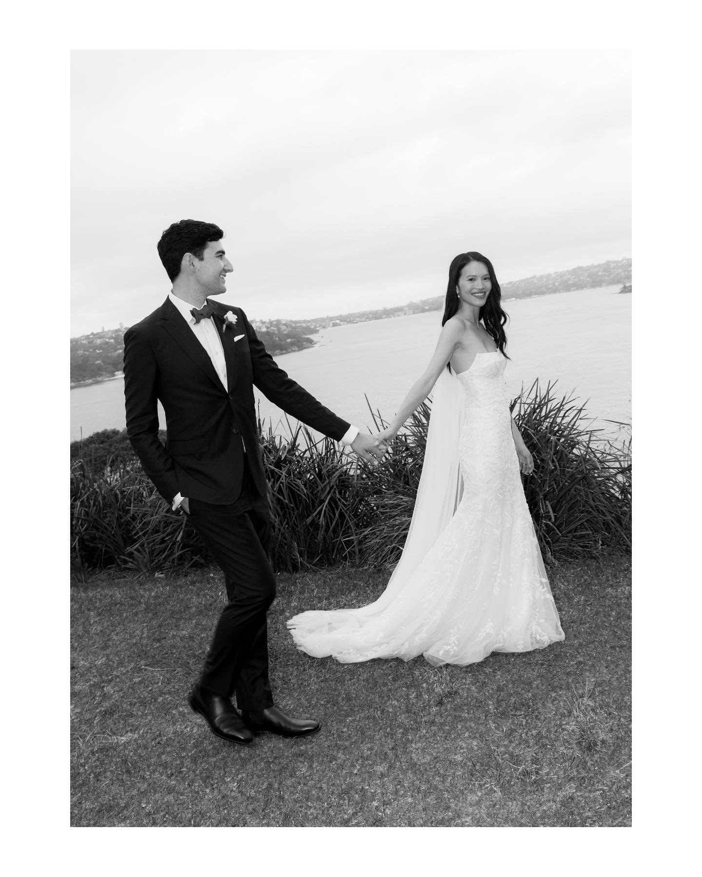 A black and white photo of a bride and groom holding hands outdoors near a body of water, with a cloudy sky above.