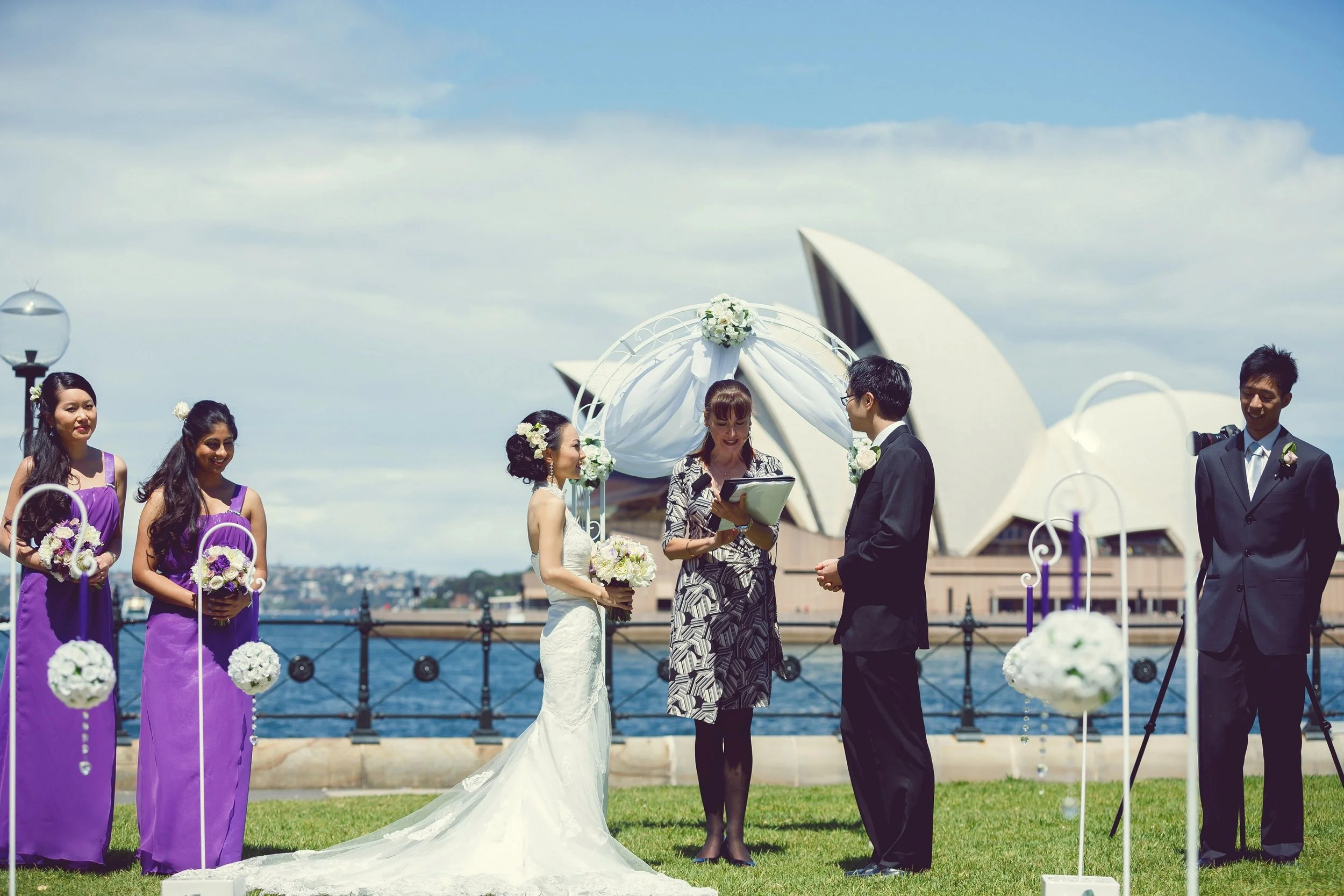 A wedding ceremony taking place outdoors near the Sydney Opera House, with the bride and groom exchanging vows, surrounded by bridesmaids and groomsmen, on a sunny day by the water.