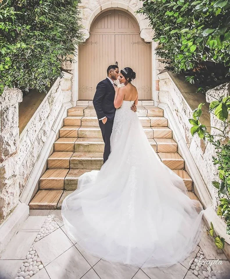 A bride and groom standing on a staircase, gazing into each other's eyes, surrounded by leafy green plants, with a closed door behind them.