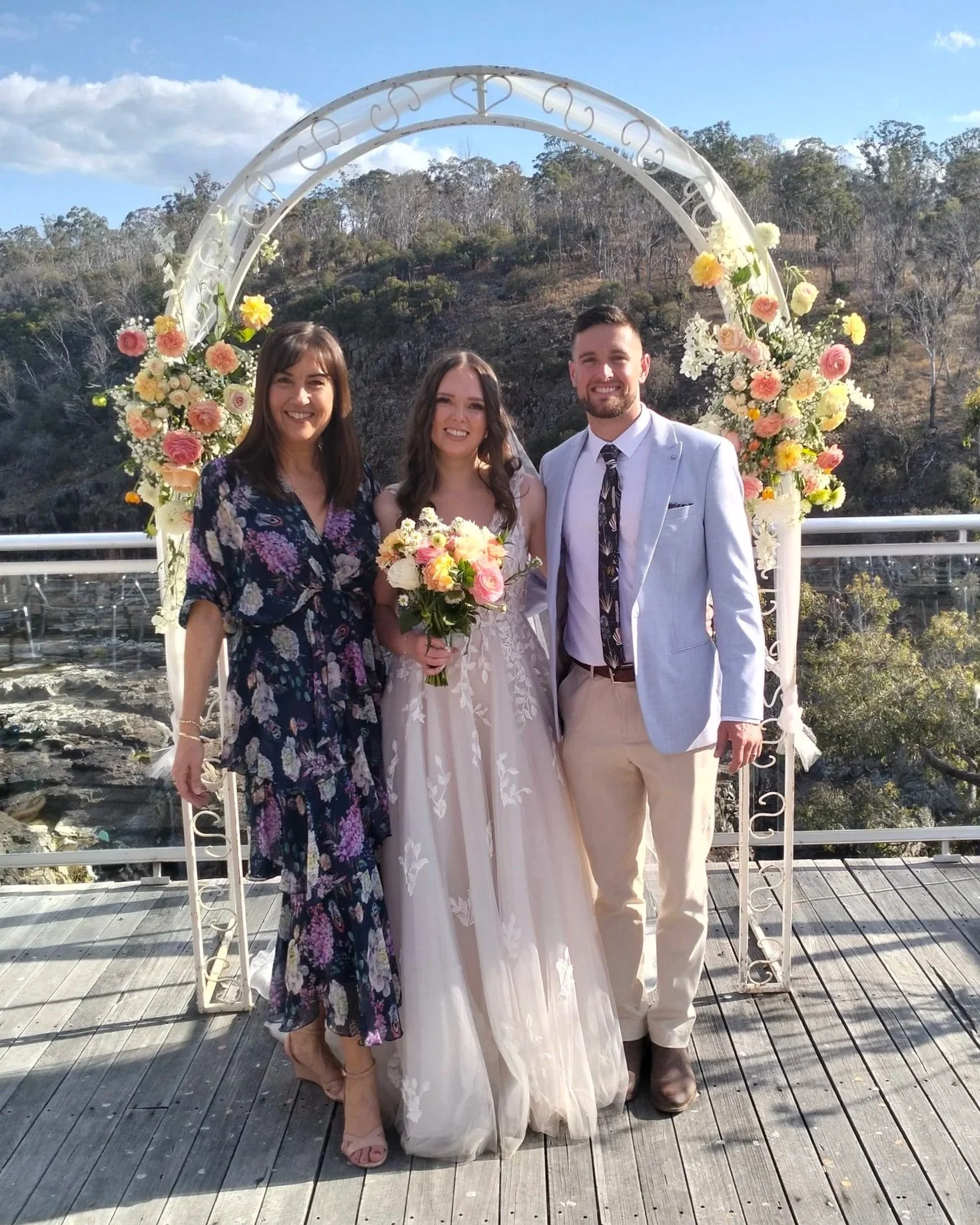 A group of three people at a wedding ceremony, standing in front of a floral arch on a wooden deck outdoors with trees and a hillside in the background.