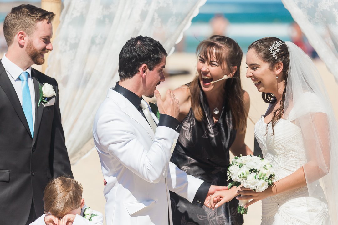 A wedding ceremony at the beach with a bride, groom, officiant, and guests smiling and holding hands.