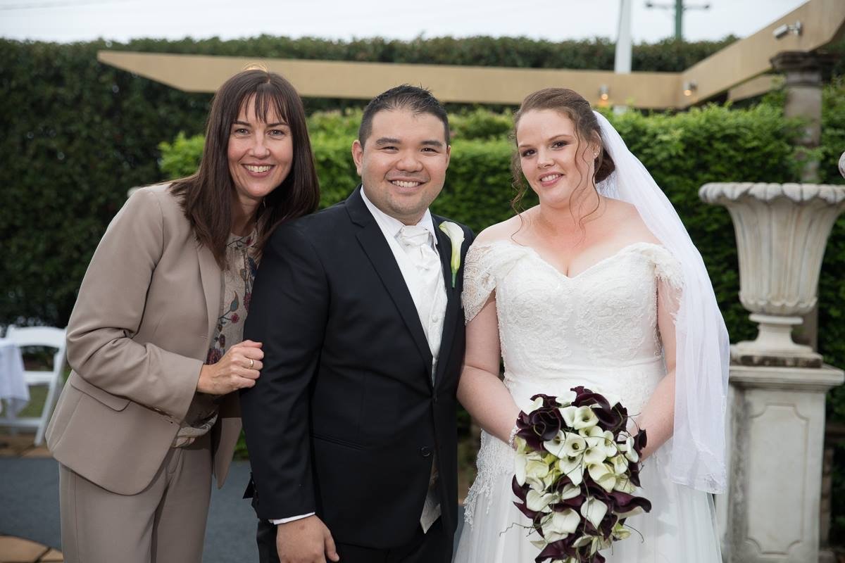 A woman, a man, and a bride standing outdoors at a wedding reception, smiling at the camera. The woman is wearing a beige suit, the man is in a black tuxedo, and the bride is in a white wedding dress holding a bouquet of dark and light calla lilies.