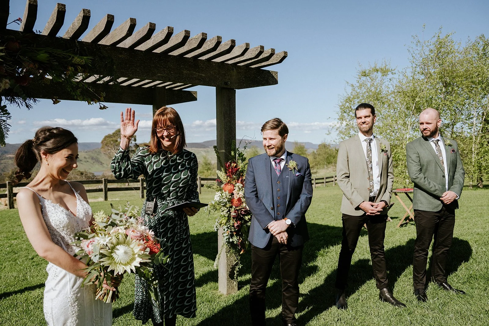 A woman in a white wedding dress holding a bouquet of flowers smiling, a woman in glasses and a patterned dress waving, three men in suits standing outside under a wooden arbor with flowers, in a grassy area with trees and mountains in the background