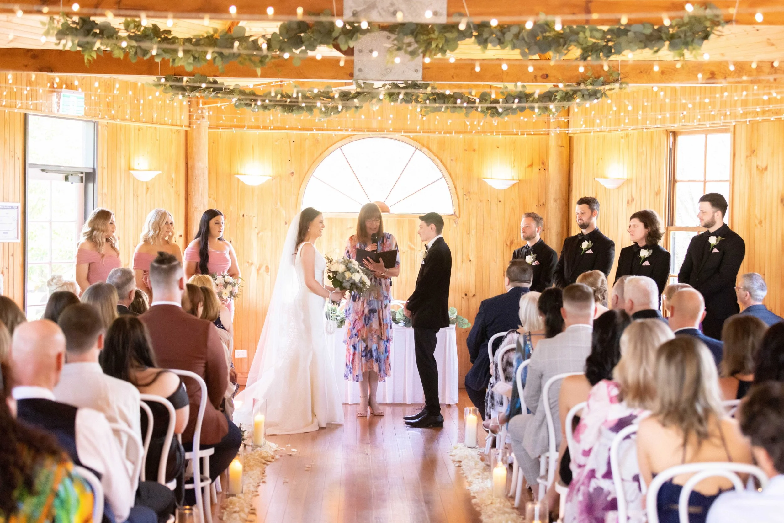 A wedding ceremony with a bride and groom exchanging vows in a rustic wooden venue decorated with string lights and greenery, with guests seated on either side and bridesmaids and groomsmen standing nearby.