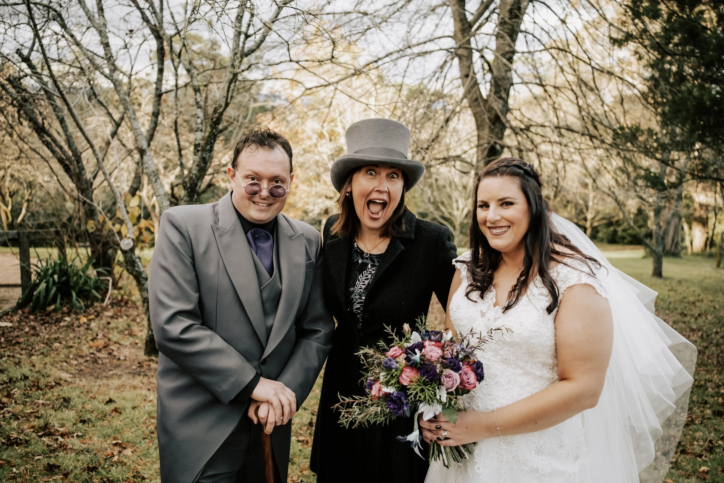 A bride in a white wedding gown holding a bouquet of purple, pink, and white flowers, standing outdoors with two people in costume, one dressed as the Mad Hatter and the other as the Queen of Hearts, all smiling and posing together.