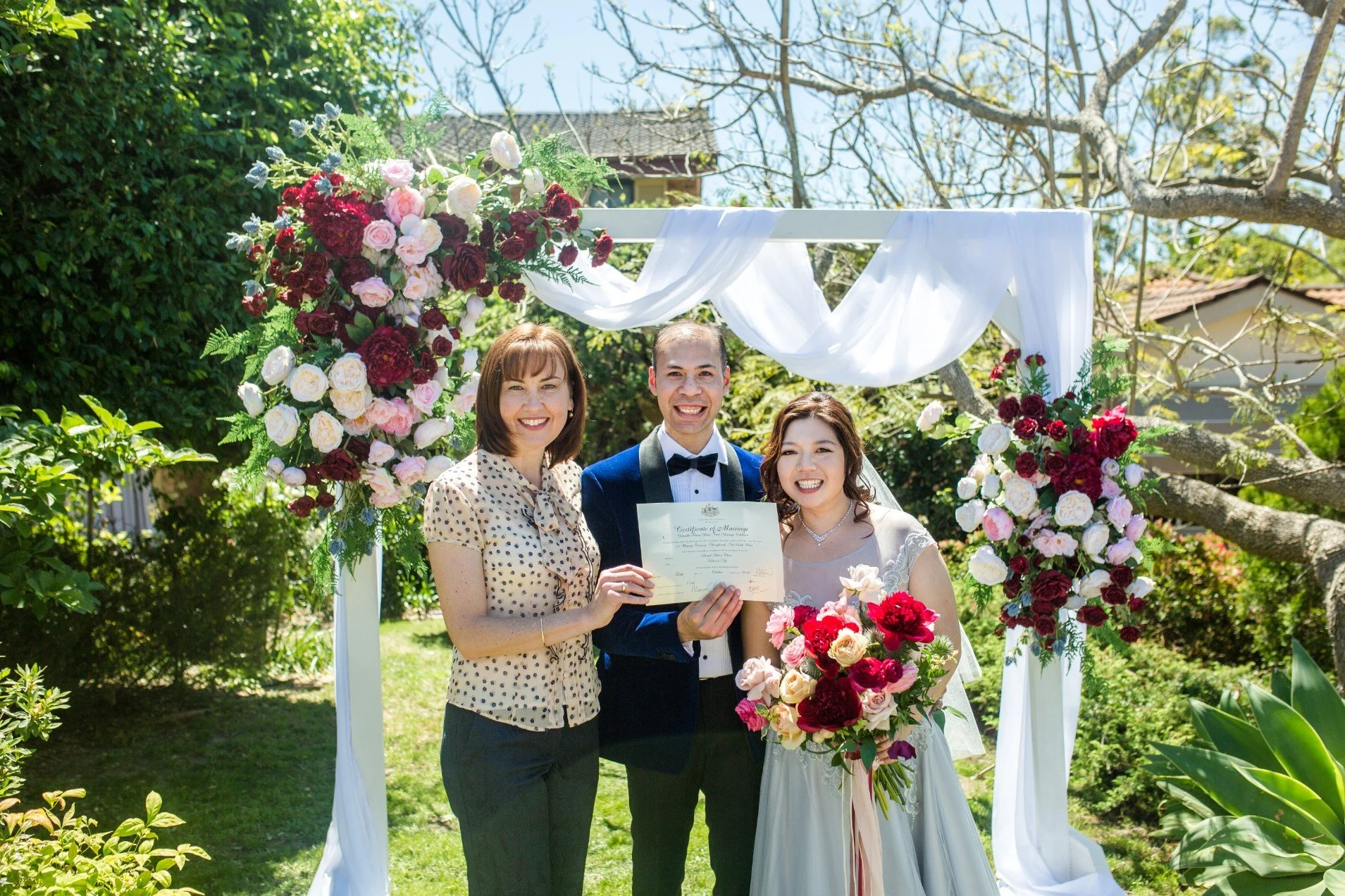 A couple gets married in a garden, holding a marriage certificate. They are smiling and standing under a floral arch with pink, red, and white flowers, with a woman standing to the left of the groom.