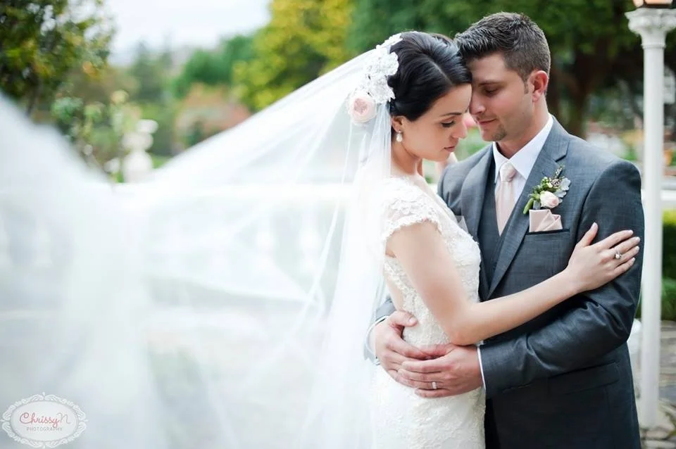 A bride and groom in a close embrace outdoors, with the bride wearing a white wedding dress and veil, and the groom in a gray suit with a boutonniere.