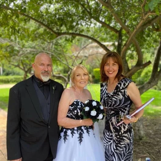 Three people standing outdoors under a tree during the daytime, including an elderly woman in a white and black dress holding a bouquet, a man in a black suit, and a woman in a black and white patterned dress holding a book or folder, smiling.