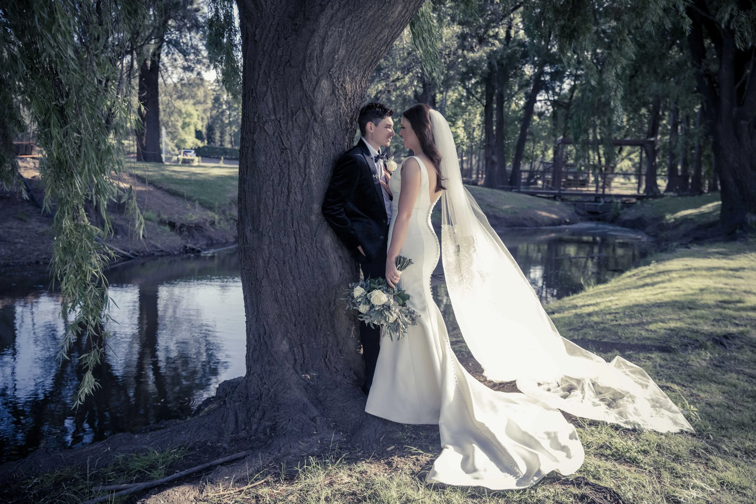 A bride and groom stand close together near a large tree by a pond in a park, gazing into each other's eyes. The bride is in a white gown with a long train and veil, holding a bouquet, while the groom is in a black tuxedo. Sunlight filters through th