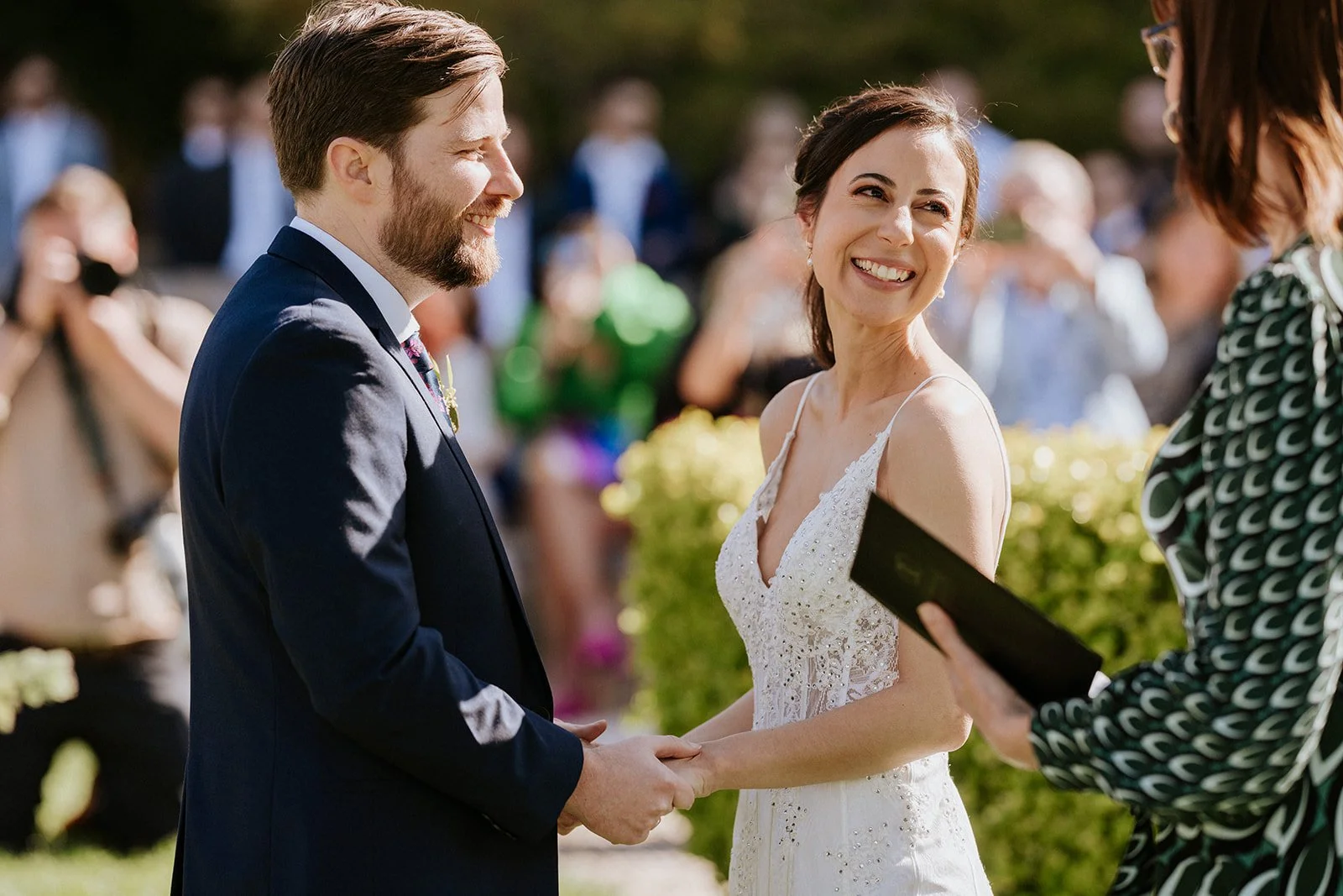A bride and groom exchanging vows during their outdoor wedding ceremony, holding hands and smiling at each other, with a officiant reading from a book in front of them and guests in the background.