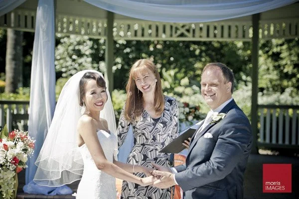 A bride and groom hold hands during their outdoor wedding ceremony, smiling at each other, with a woman officiant in the background. They are under a decorated gazebo, surrounded by greenery and flowers.