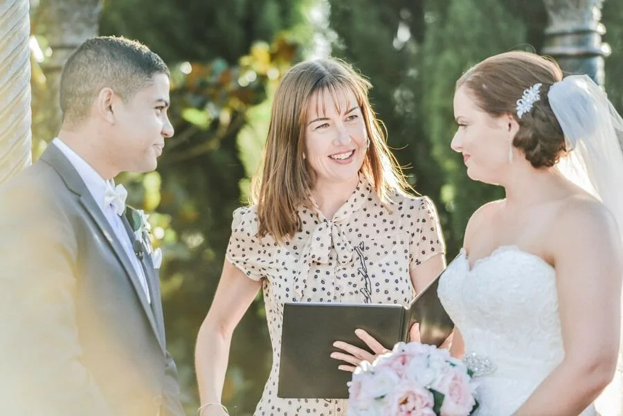 A bride and groom exchanging vows outdoors during their wedding ceremony, with a smiling officiant holding a book between them.