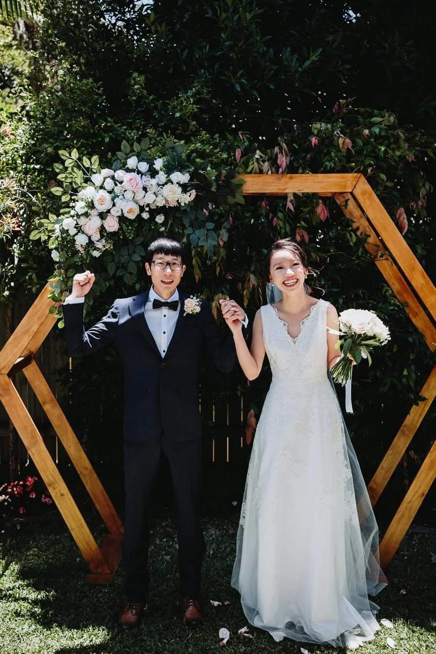 A bride and groom celebrating outdoors in front of a floral arch and wooden geometric structure, with the groom in a tuxedo and the bride in a white wedding dress holding a bouquet.