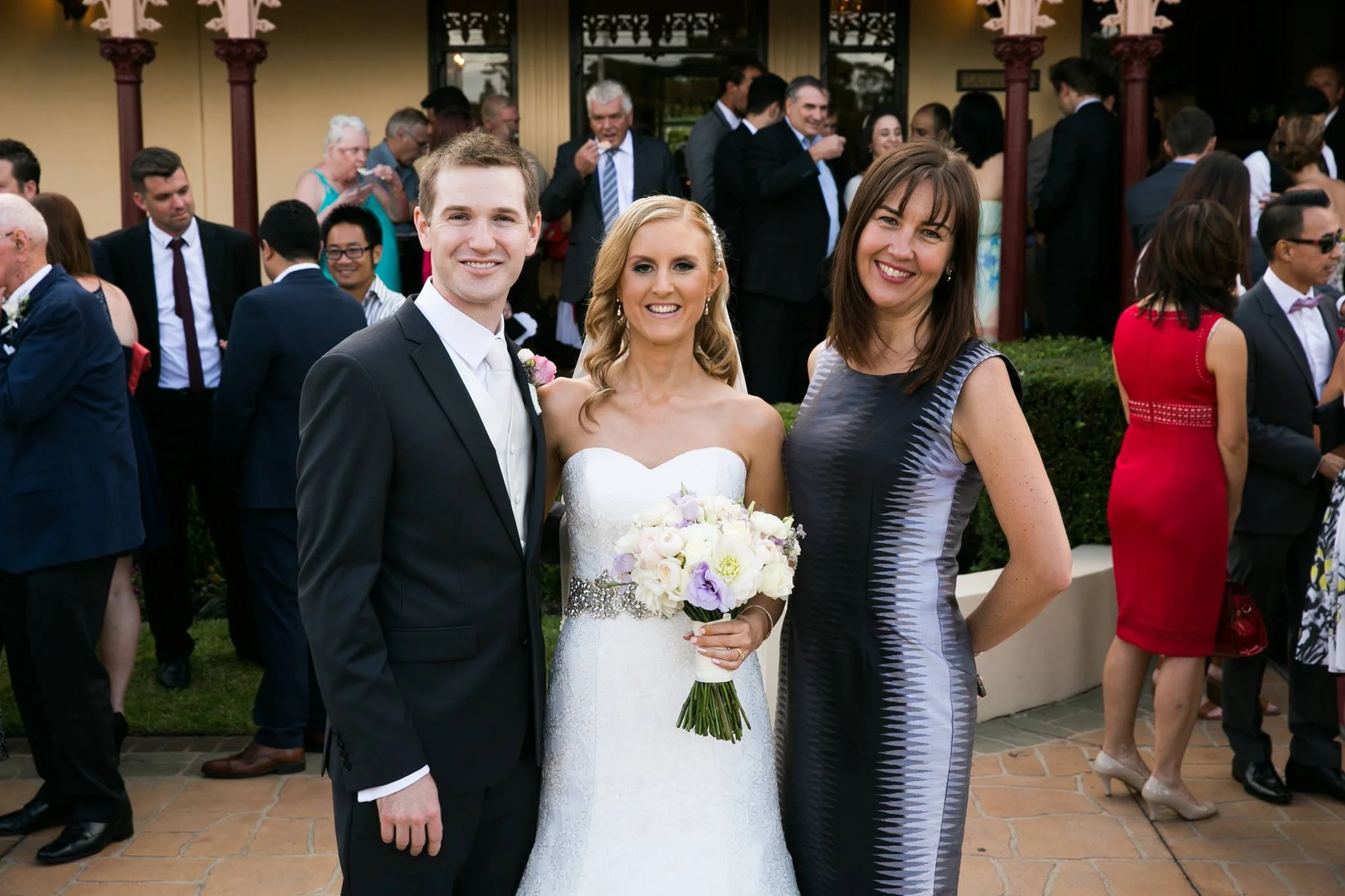 Three people, a groom, bride, and woman, smiling for a photo at a wedding reception outdoors with many guests in the background.