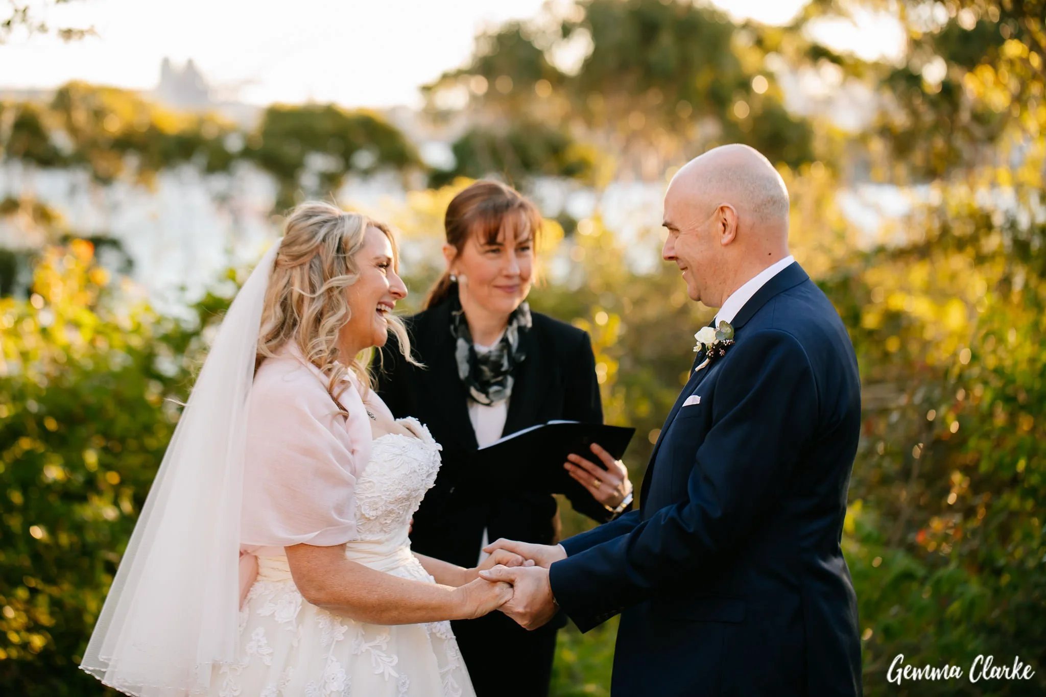 A bride and groom holding hands and smiling at each other during a wedding ceremony outdoors, with an officiant standing behind them and holding a notebook, surrounded by greenery and trees.