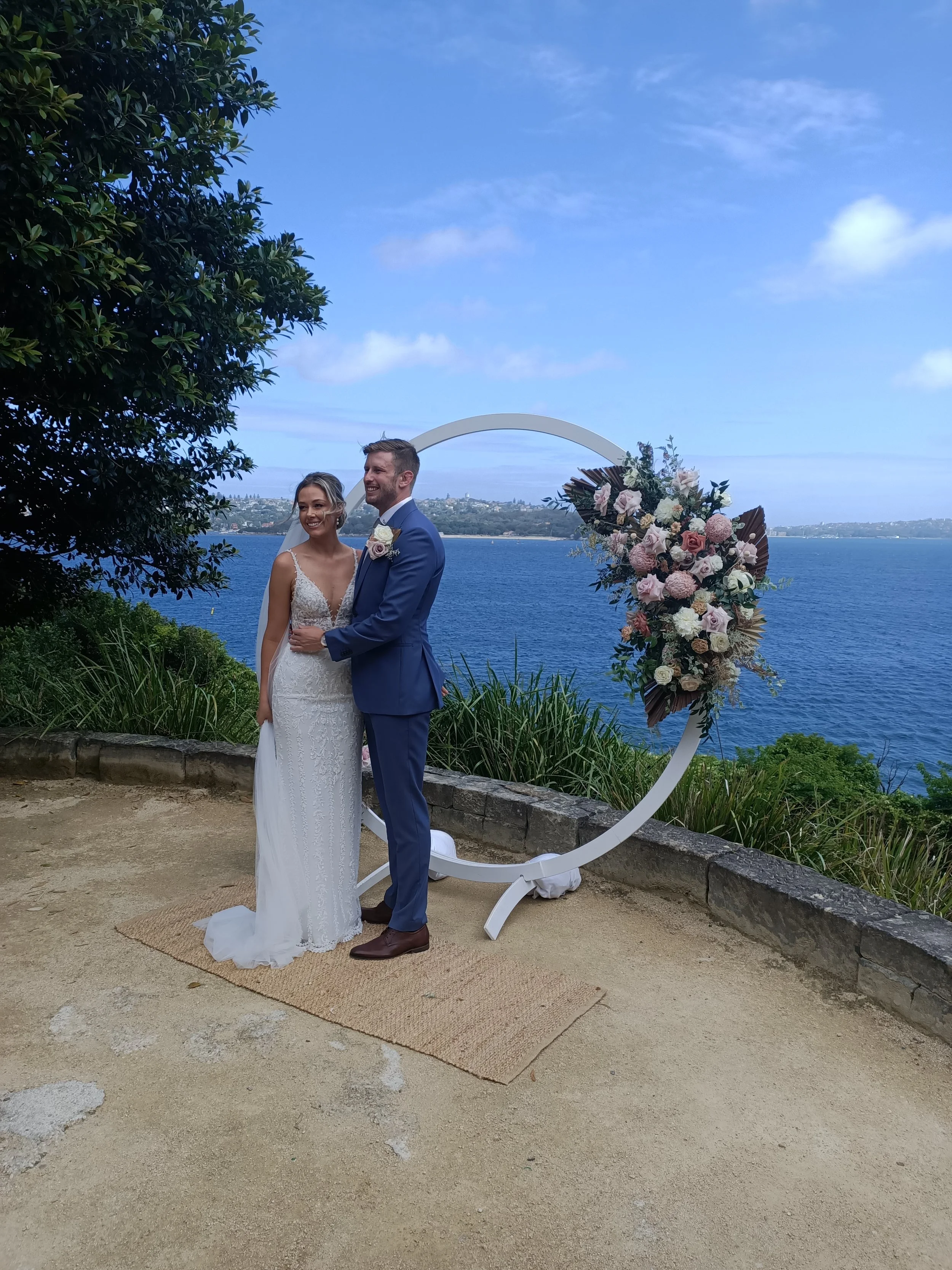 A bride and groom standing together outdoors near a body of water, with a decorative floral arch behind them.