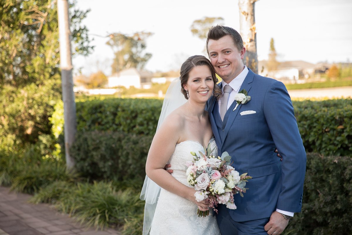A newlywed couple smiling outdoors on a sunny day, the bride in a wedding dress holding a bouquet, and the groom in a blue suit with a white boutonniere.