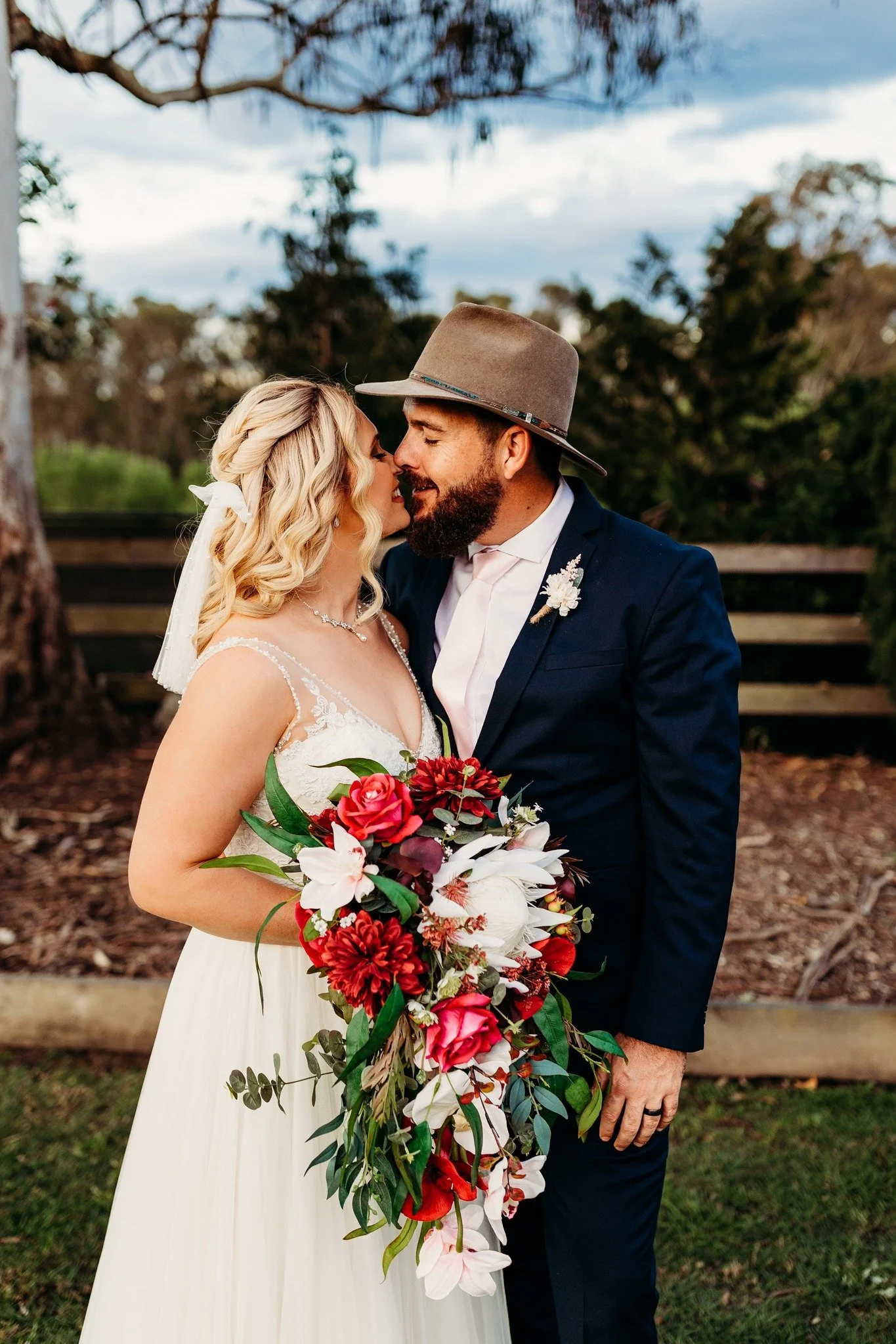 A bride and groom sharing a moment outdoors on their wedding day, with the bride holding a large bouquet of red and white flowers and greenery, and the groom wearing a gray hat and navy suit.