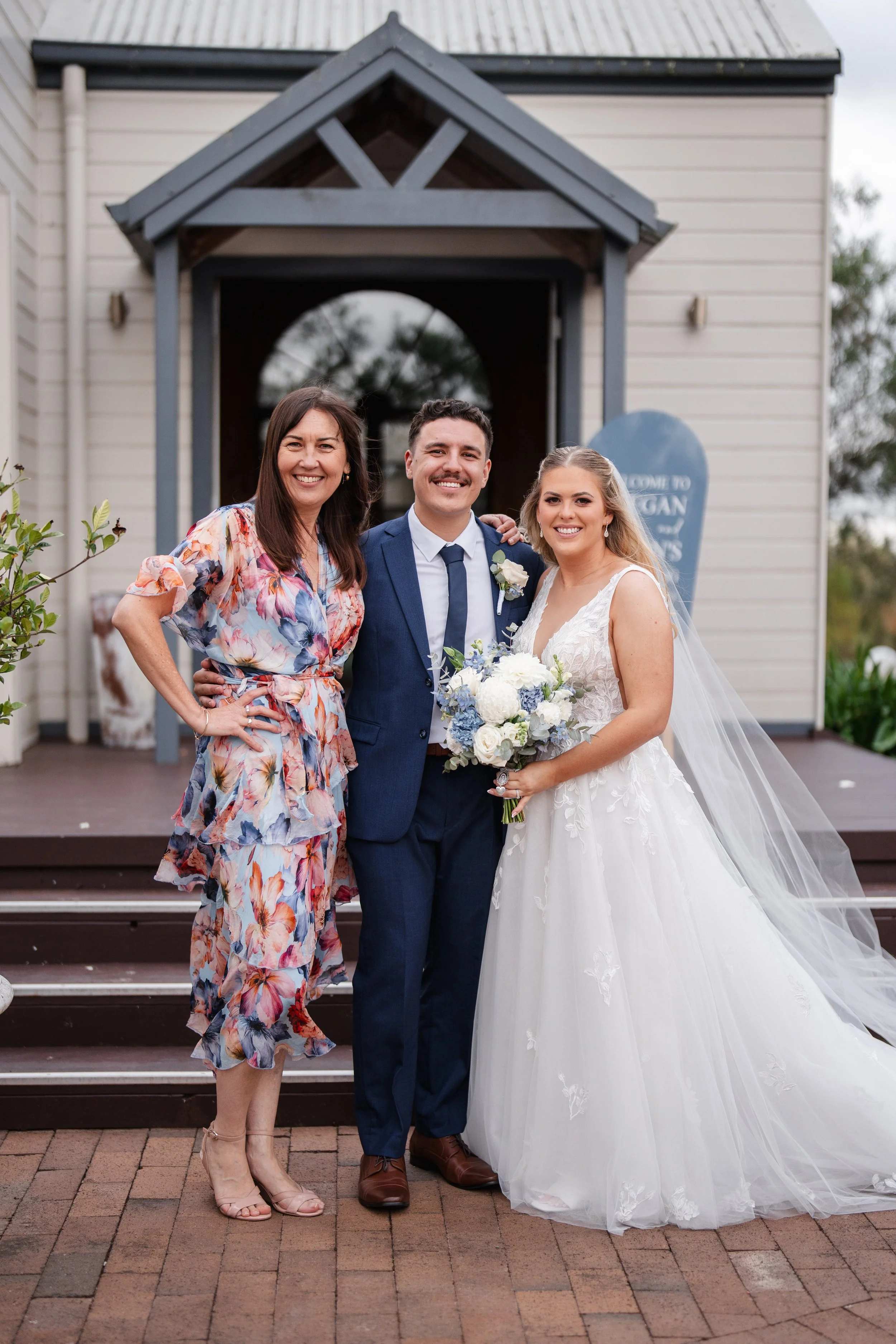 A bride, groom, and woman standing outside on stairs in front of a building with a sign, smiling for the camera.