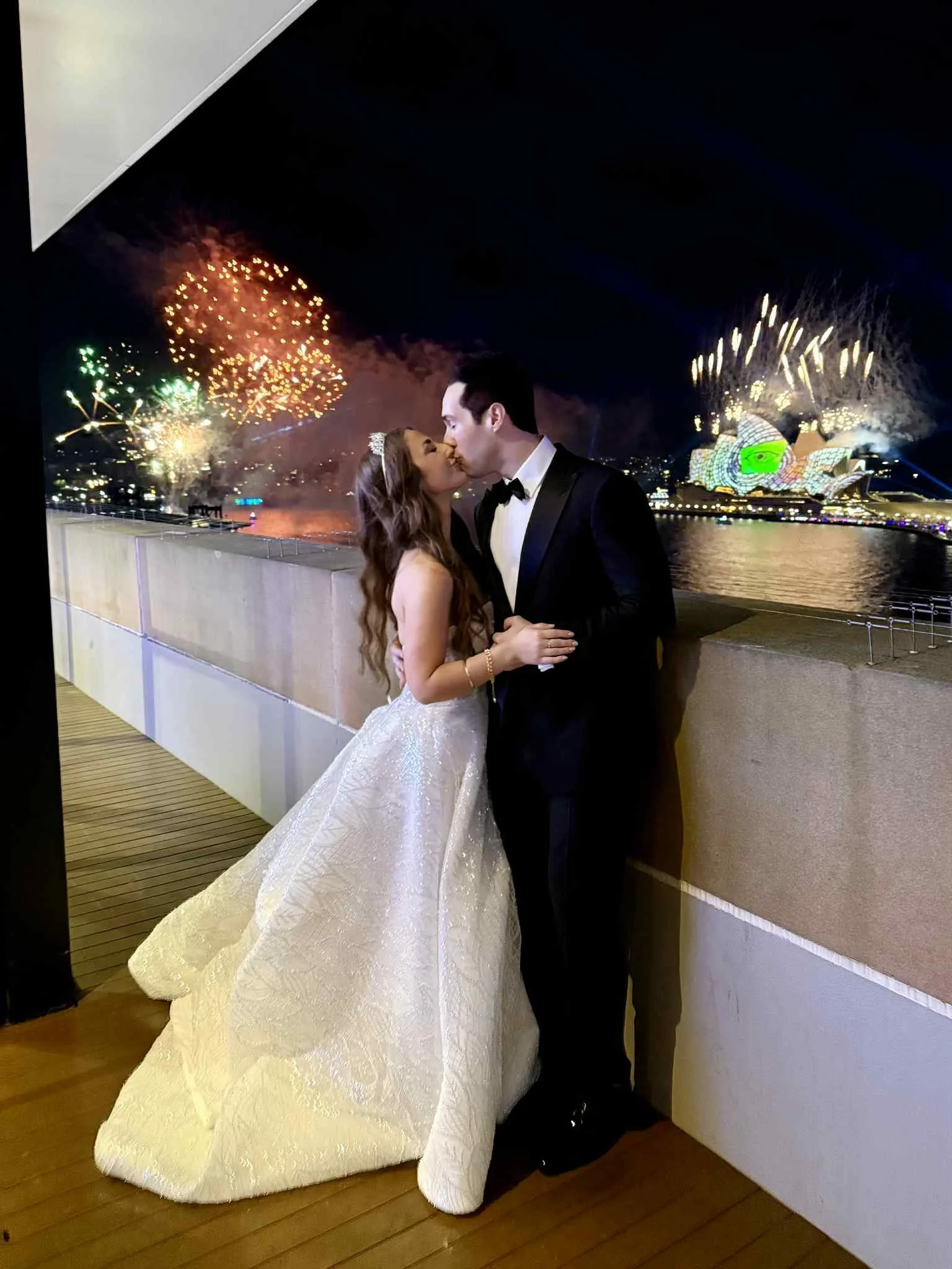 A newlywed couple sharing a kiss on a balcony at night, with fireworks over the Sydney Opera House and Harbour Bridge in the background.