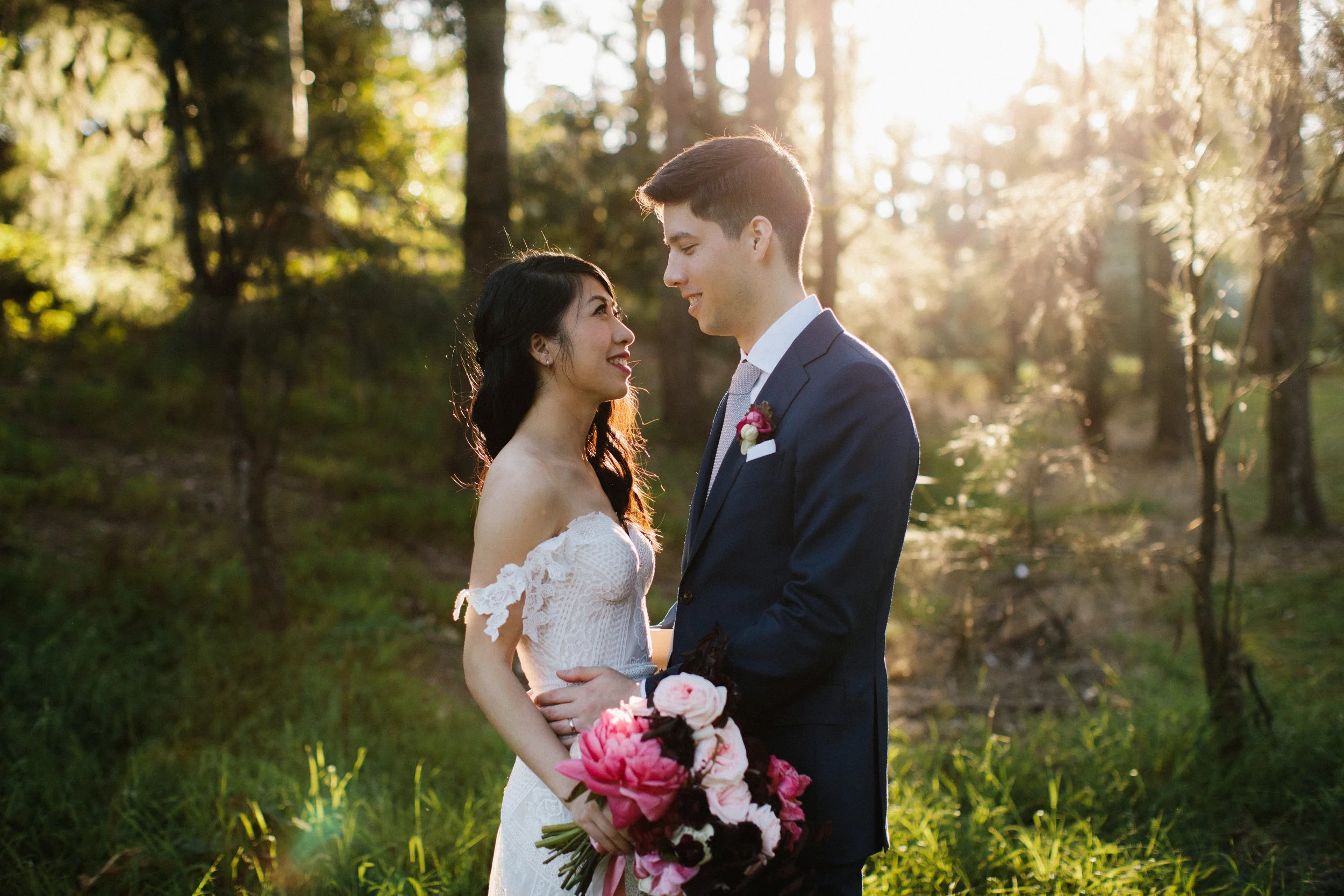 A bride and groom standing in a sunlit forest, gazing into each other's eyes. The bride is holding a bouquet of pink and white flowers, wearing an off-shoulder white wedding dress. The groom is dressed in a navy suit with a boutonniere. Sunlight filt