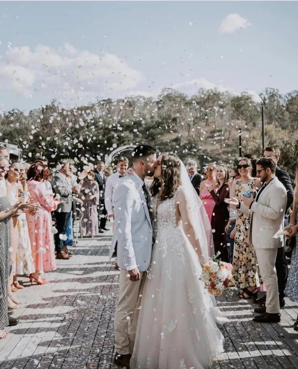 A bride and groom share a kiss at their outdoor wedding ceremony as guests throw confetti in the air.