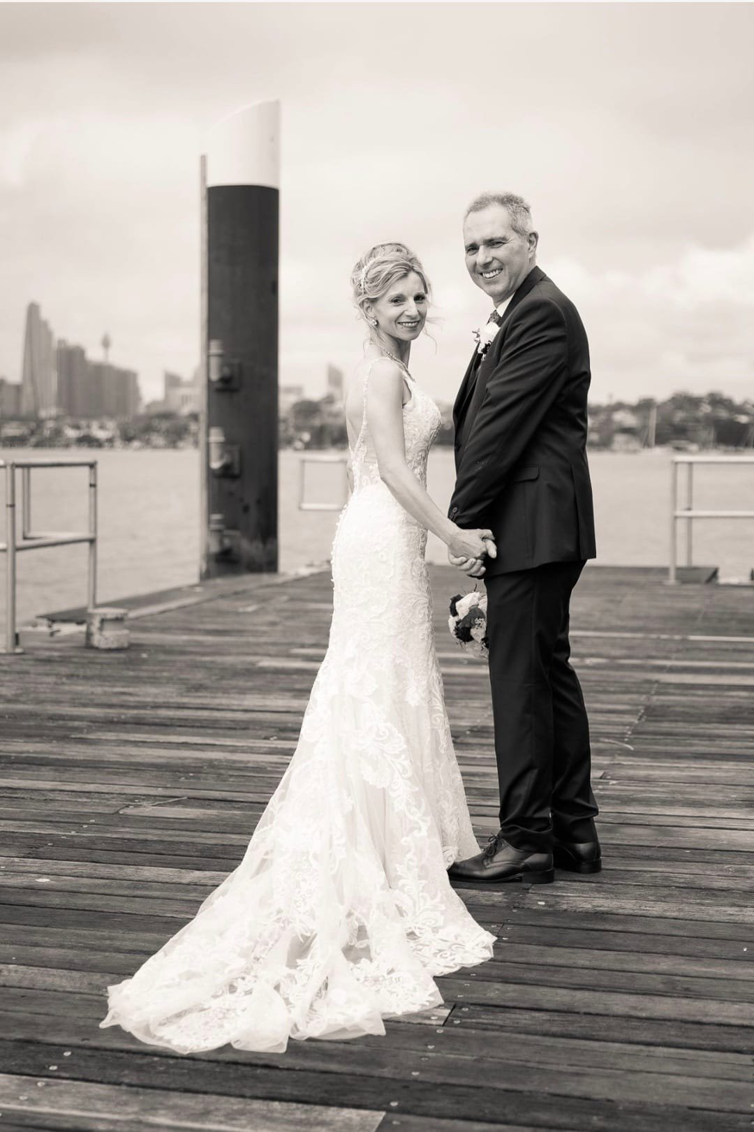 Black and white photo of a newly married couple standing on a wooden dock, holding hands and looking at the camera with city skyline and water in the background.