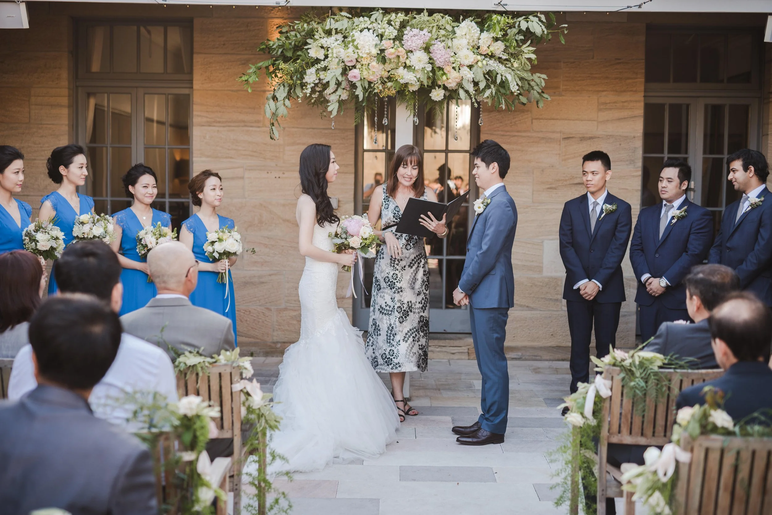 A wedding ceremony with a bride and groom exchanging vows outdoors, surrounded by bridesmaids in blue dresses and groomsmen in dark suits, with guests seated watching.