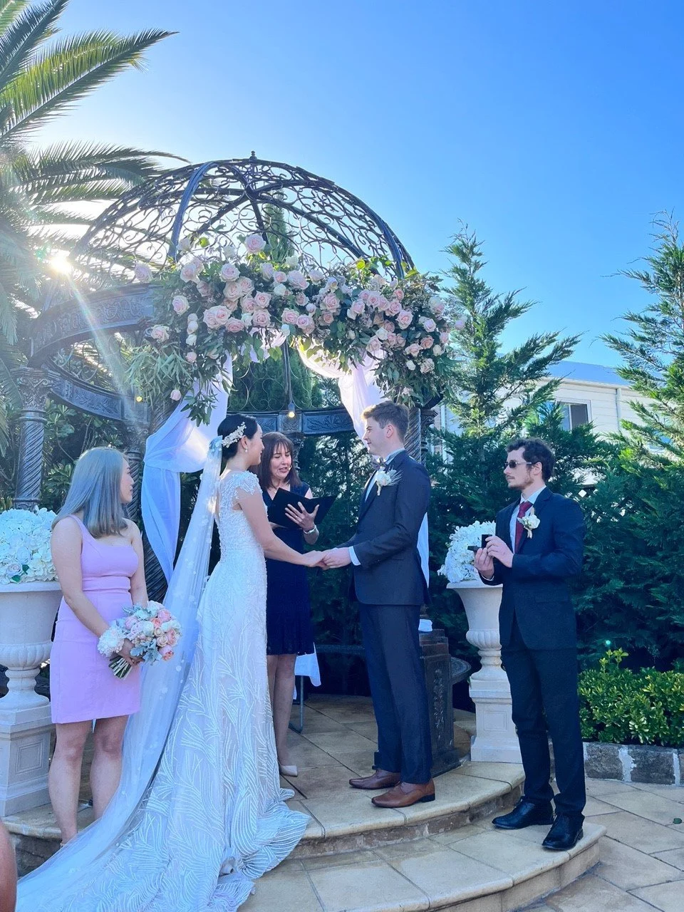 A wedding ceremony outdoors with a bride and groom holding hands under a floral arch, surrounded by bridesmaids and groomsmen, in a garden setting with blue sky and palm trees.