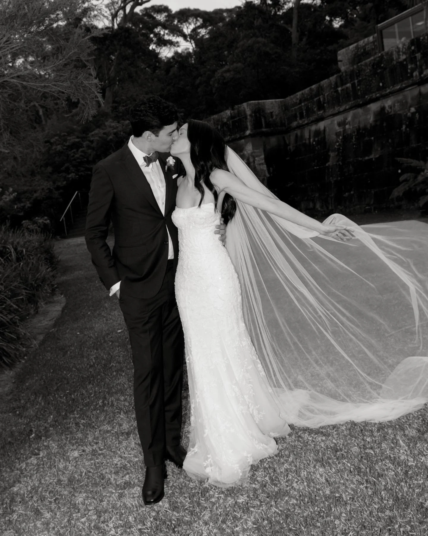 A black and white photograph of a newlywed couple kissing outdoors. The groom is in a black tuxedo with a bow tie, and the bride is in a strapless lace wedding gown with a long veil. The bride is extending her arm behind her, and they are standing on