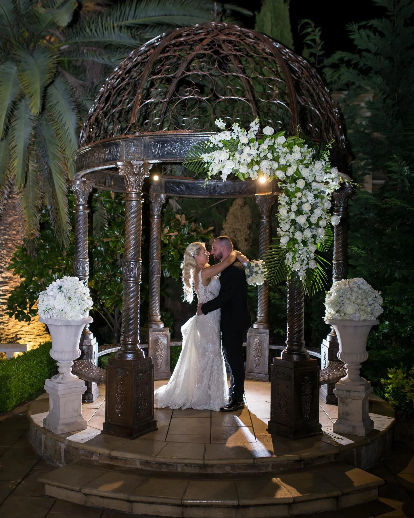 A bride and groom are embracing in an outdoor wedding setting under a decorative metal gazebo decorated with white flowers and greenery. The scene is illuminated at night surrounded by lush greenery.
