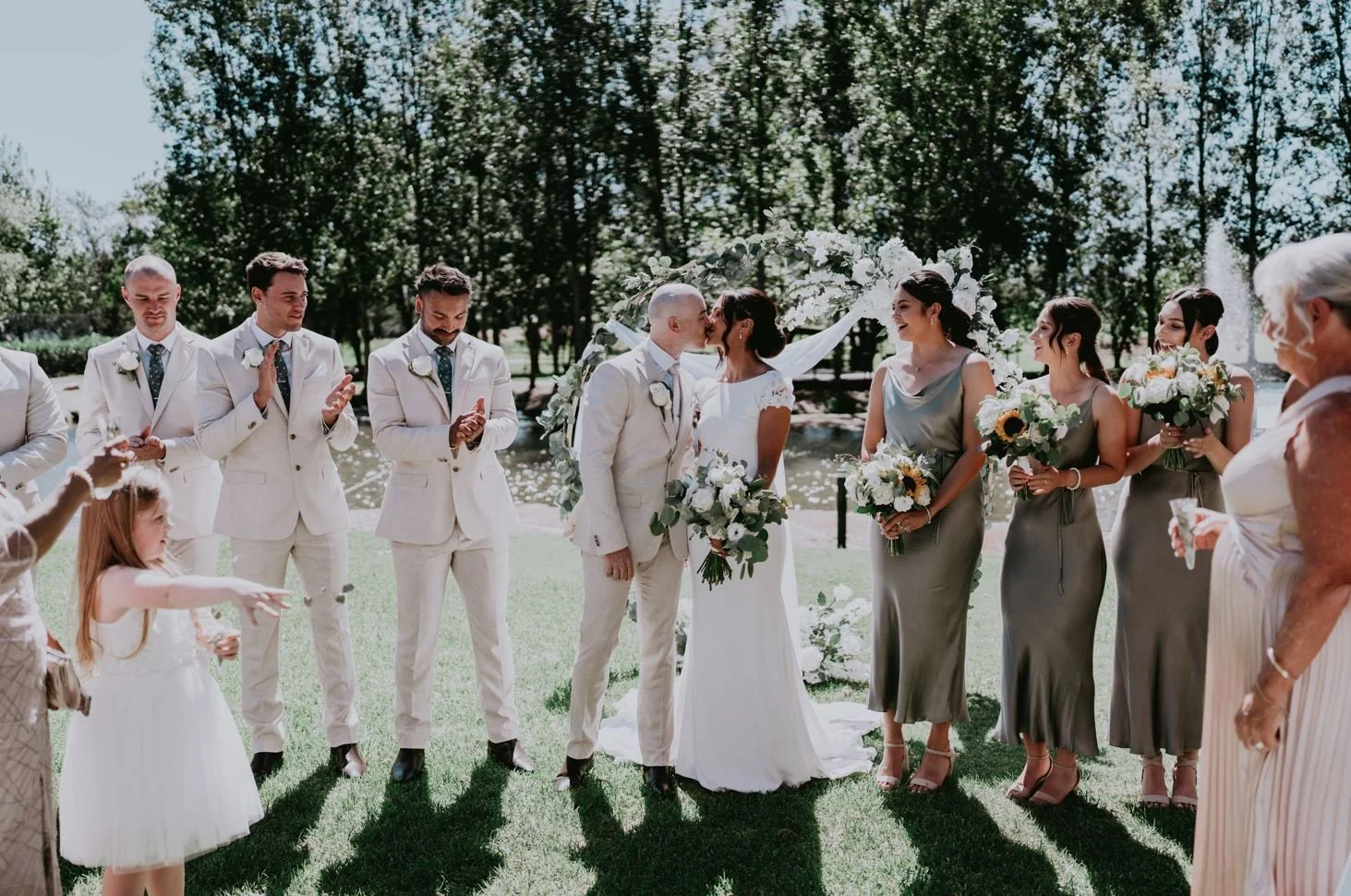 Bride and groom kissing during outdoor wedding ceremony with bridesmaids and groomsmen clapping, set in a park near a lake, on a sunny day.