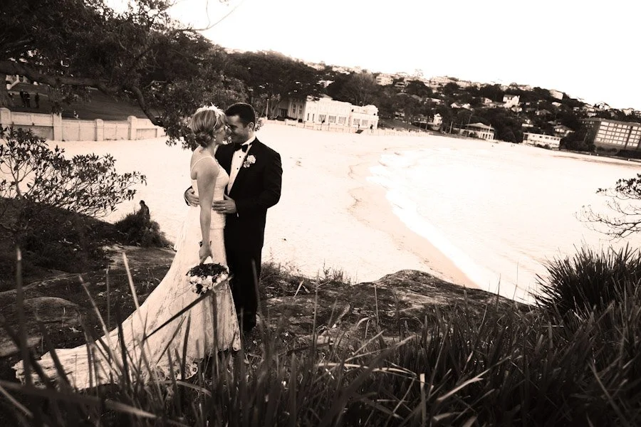 A bride and groom sharing a kiss on a grassy hill overlooking a beach and ocean at sunset, dressed in wedding attire, with the bride in a white gown holding a bouquet.