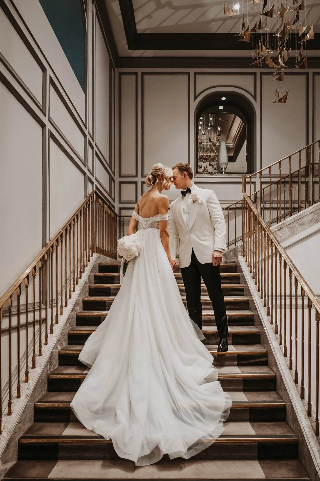 A bride and groom in wedding attire on a staircase, facing each other with foreheads touching, in an elegant indoor setting.