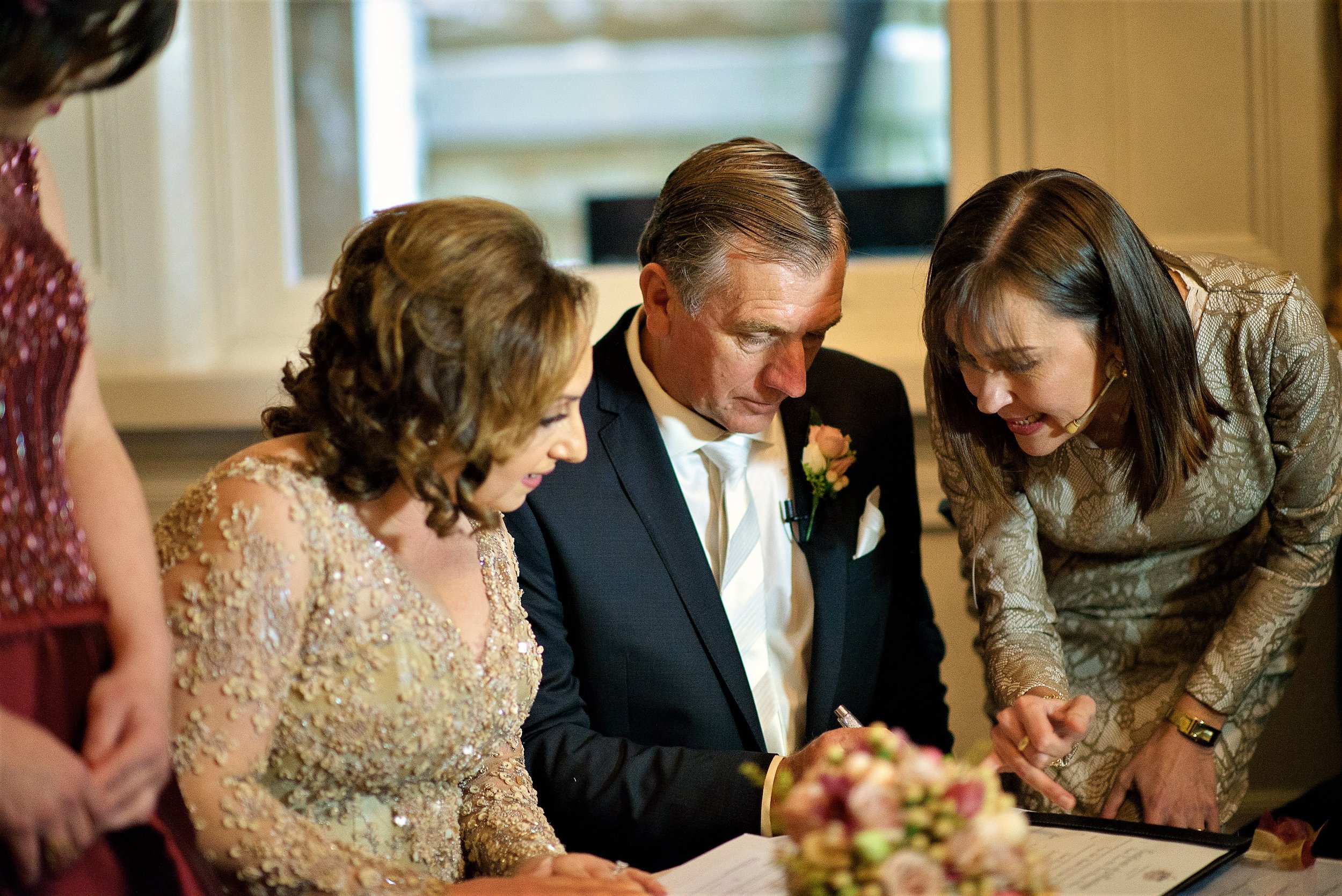 A wedding ceremony with four people gathered around a signing book or document, including a groom in a dark suit and woman in a gold lace dress, with two other women in elegant dresses leaning in for a closer look. They appear to be signing a marriag