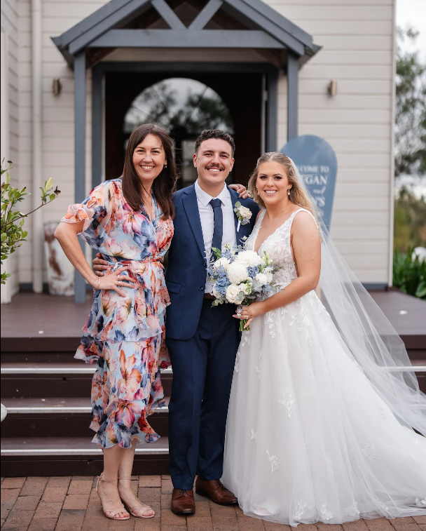 A bride, groom, and a woman (possibly family or friend) standing together on a wedding day in front of a house, with the bride holding a bouquet of white and blue flowers, all smiling for the photo.