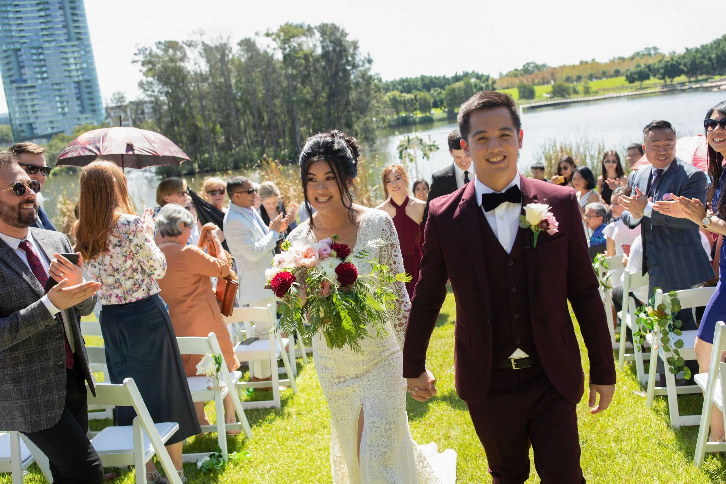A newlywed couple walks hand-in-hand down the aisle outdoors by a lake, smiling, with friends and family applauding and taking photos.