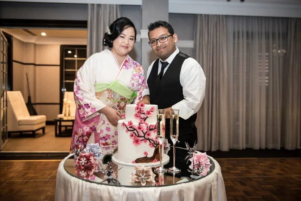 A woman in a kimono and a man in formal attire stand behind a decorated wedding cake with pink flowers, champagne flutes, and floral decorations on a table in a well-lit room.