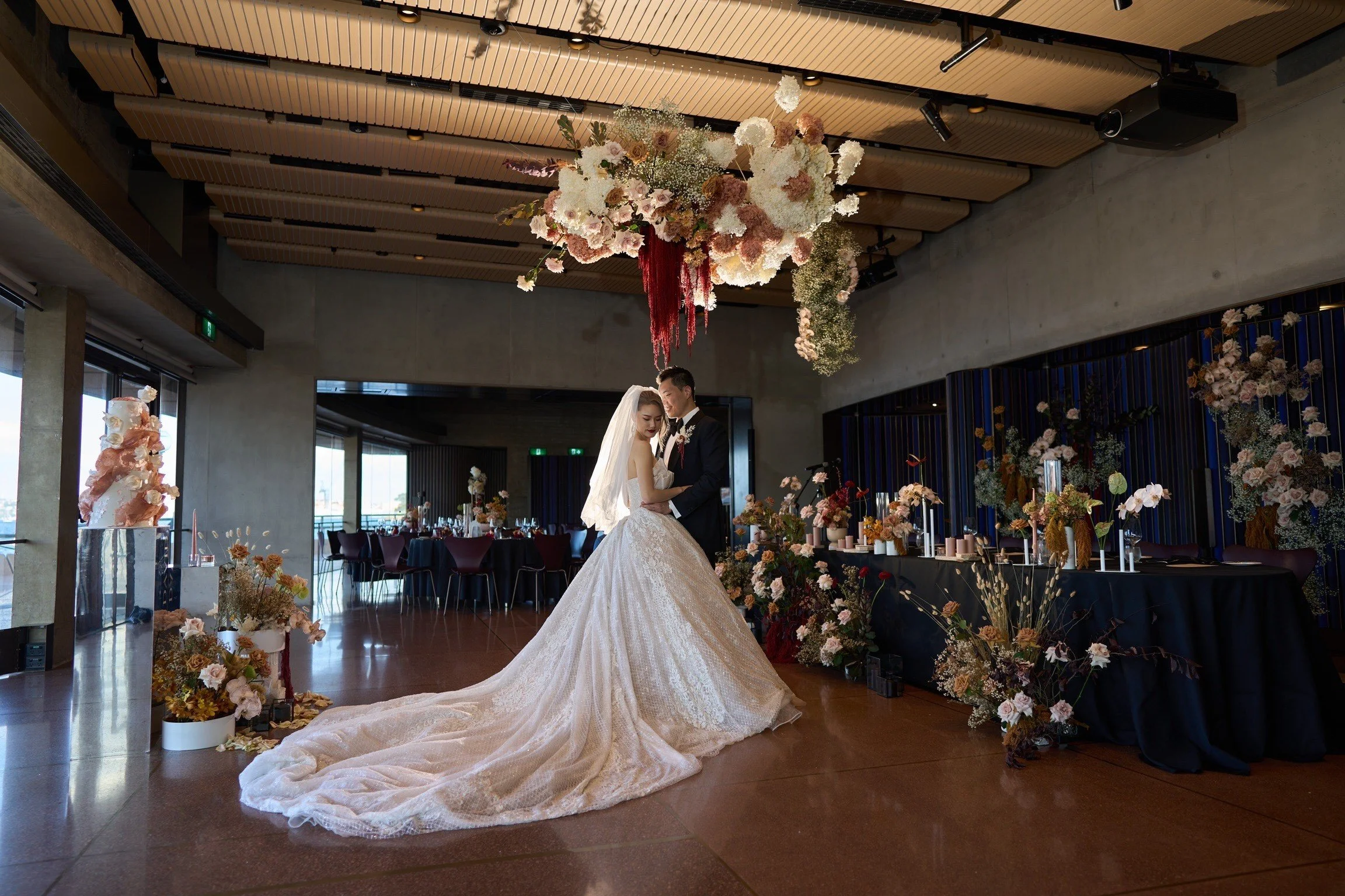 A bride and groom sharing a dance in a wedding reception hall decorated with floral arrangements and a large floral ceiling installation.