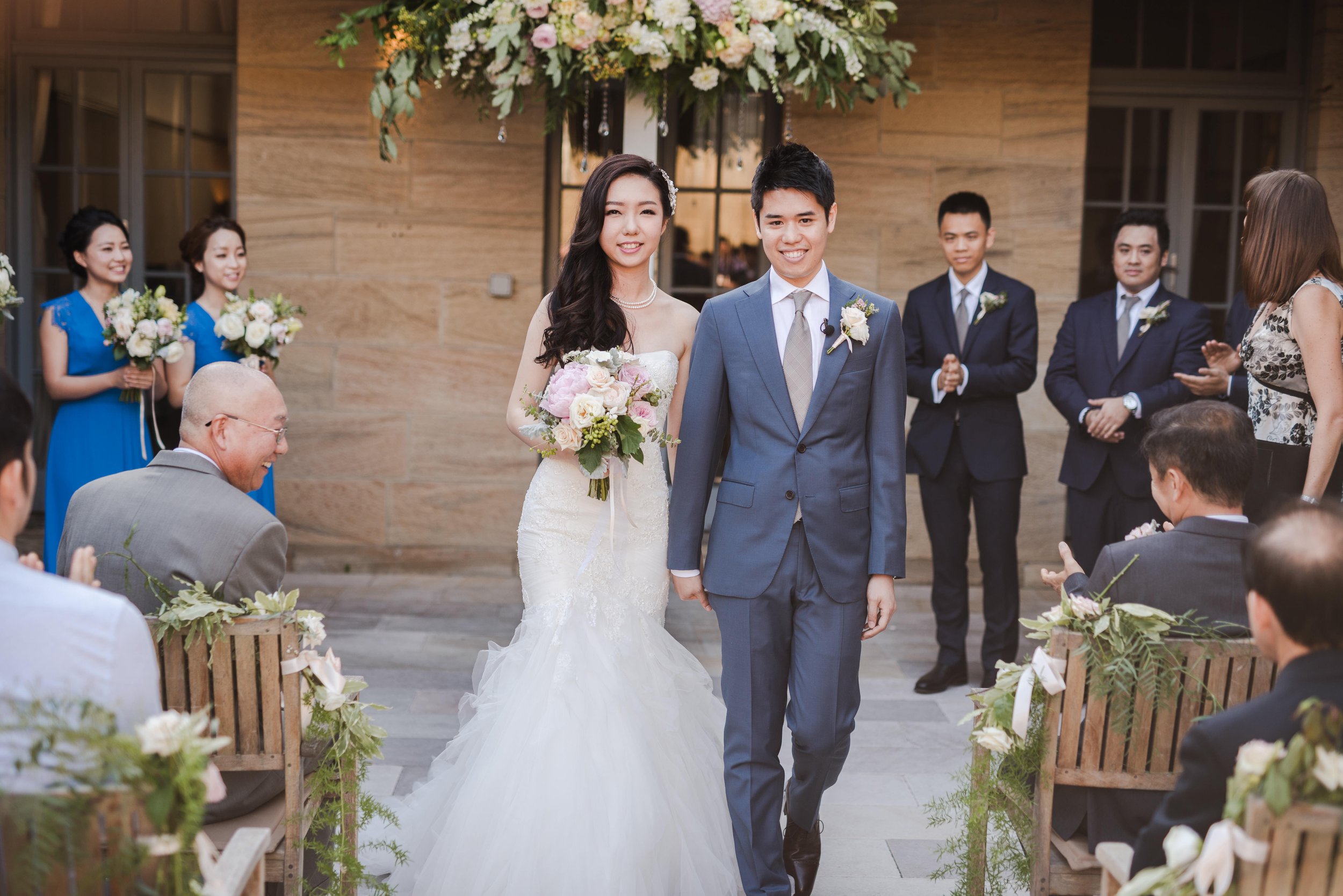 A bride and groom walk down the aisle at their wedding ceremony. The bride is wearing a white gown and holding a bouquet of pink and white flowers. The groom is in a blue suit with a white shirt and gray tie. Guests and bridesmaids in blue dresses an