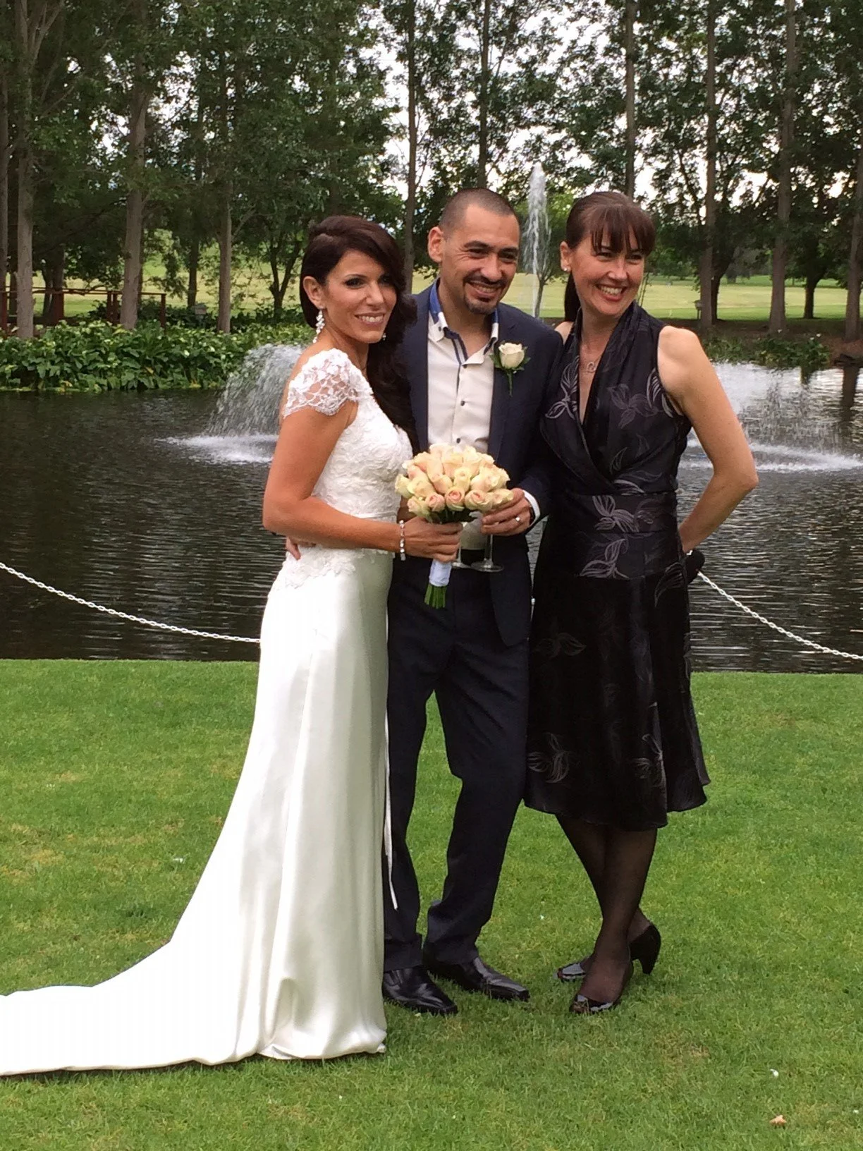 A bride, groom, and a woman standing by a pond with a fountain in the background, outdoors during a wedding celebration.