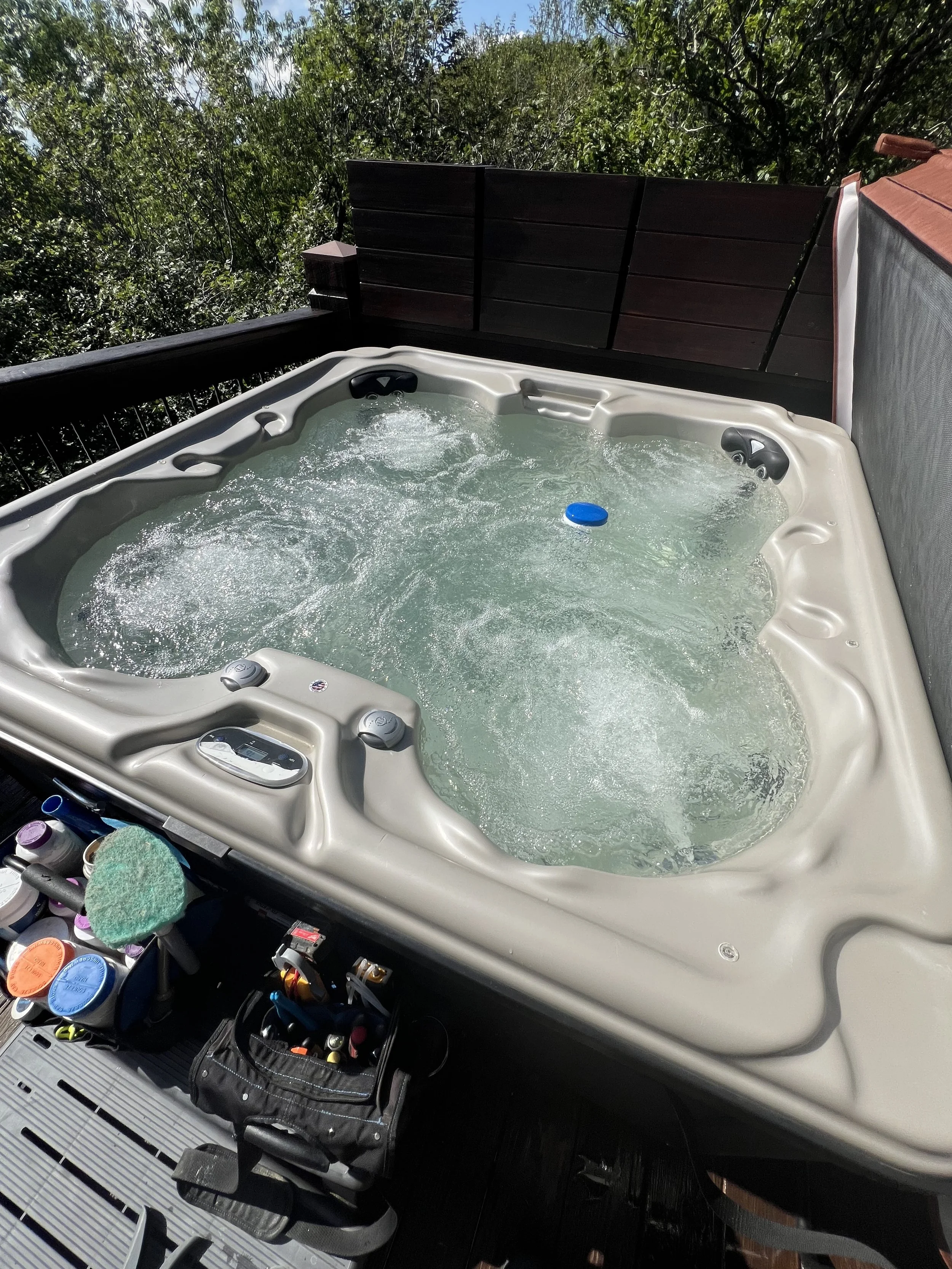 Hot tub on outdoor deck surrounded by greenery, with a wooden privacy fence and spa accessories nearby.